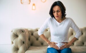 Adult woman is sitting on a sofa at home and touching her lower stomach while suffering from cramps.