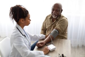 Hypertension In Older Age. Young Female Medical Worker Measuring Arterial Blood Pressure Of Senior Black Man Using Cuff, Patient Having Problems With Tension, Sitting At Table. Health Care Concept