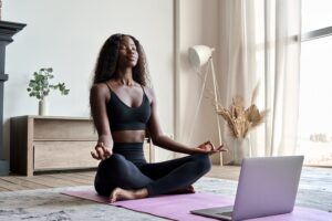 Young calm fit healthy African black woman sitting on floor at home doing yoga breathing exercise, meditating learning online training virtual class on computer. Exercises for mental health concept.