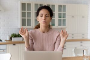 Home office yoga. Calm millennial hispanic female freelancer sit by pc with closed eyes breath deep meditate. Mindful young woman relax from online work doing breathing exercises join fingers in mudra