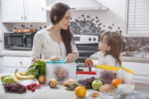 A young mother puts a sandwich into a clear food storage bag. Her daughter is handing her a peach. They are in a bright modern kitchen with a concrete counter top surrounded by other fruits and vegetables that she is preparing and storing.